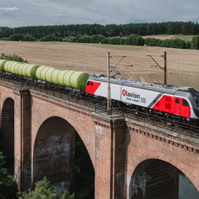 Pociąg z lokomotywą elektryczną i wagonami na torze. Logo GRUPA & UNIMOT znajduje się na boku wagonu kolejowego. Olavion ESACT056.